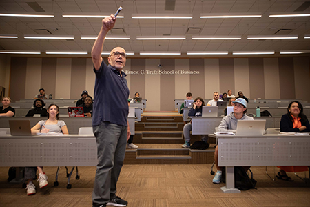 An instructor points towards the camera with students in a lecture hall behind him.